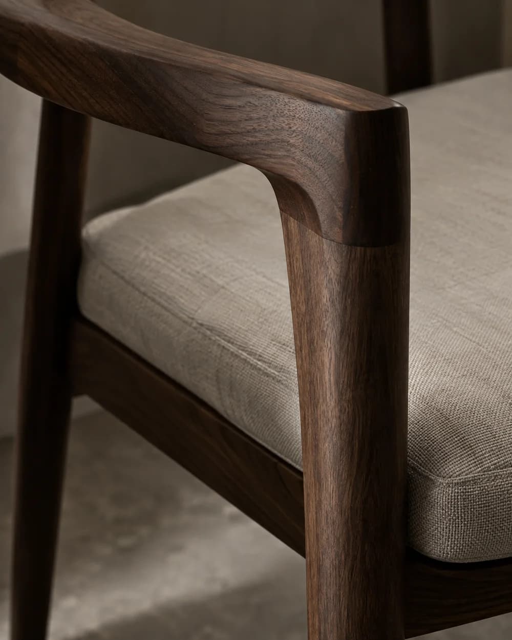 Concept detail of a curved walnut chair arm meeting its leg beside a woven linen cushion.